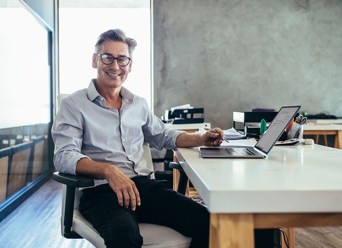 Business Insurance - Smiling Business MAn Sitting at Desk in a Modern Office