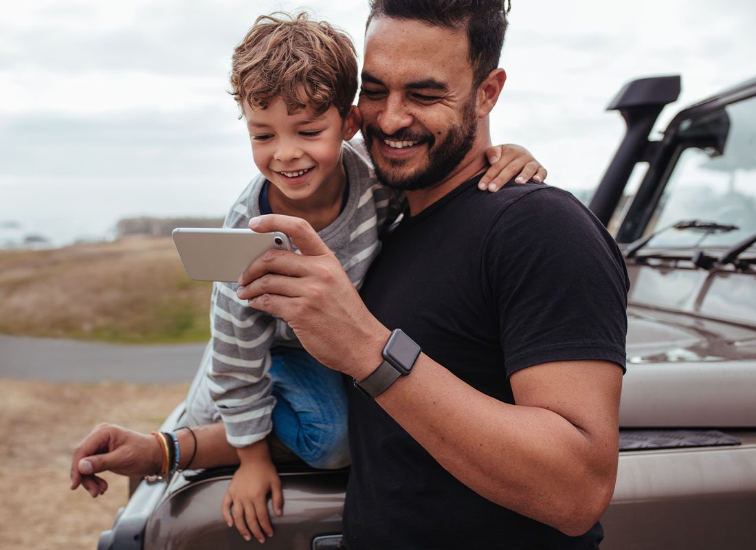 Contact - Smiling Father and Son Holding a Phone and Leaning Against a Car Outdoors